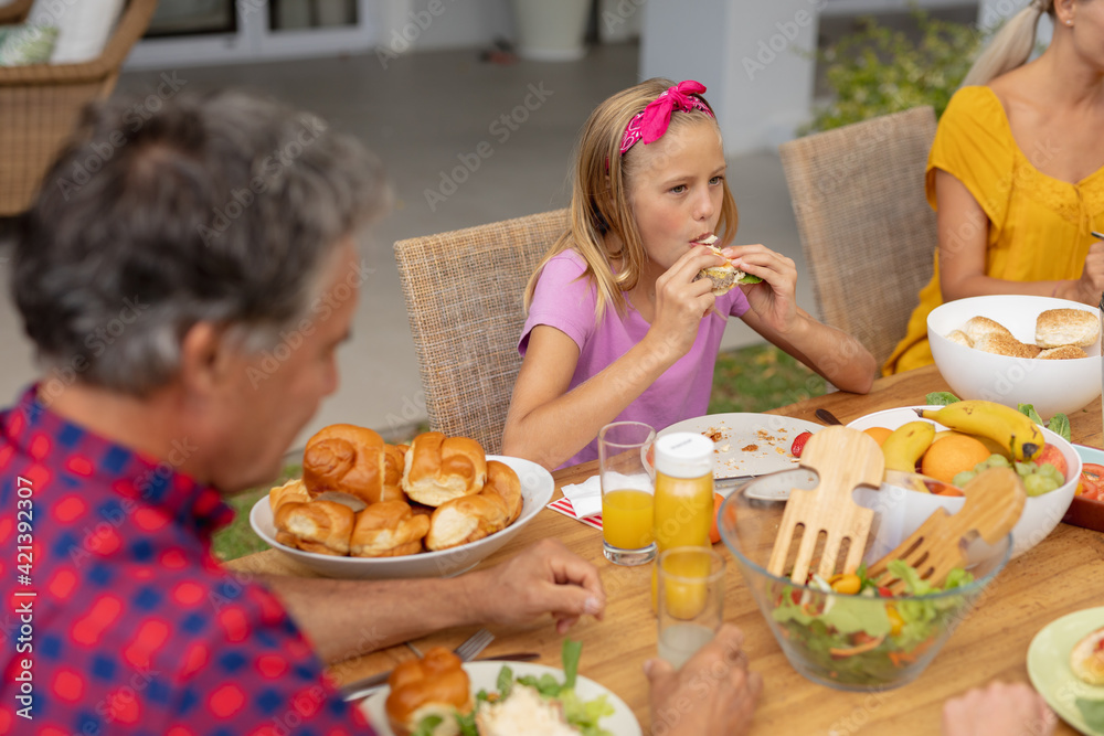 Caucasian girl eating hamburger sitting at table with family having meal in garden