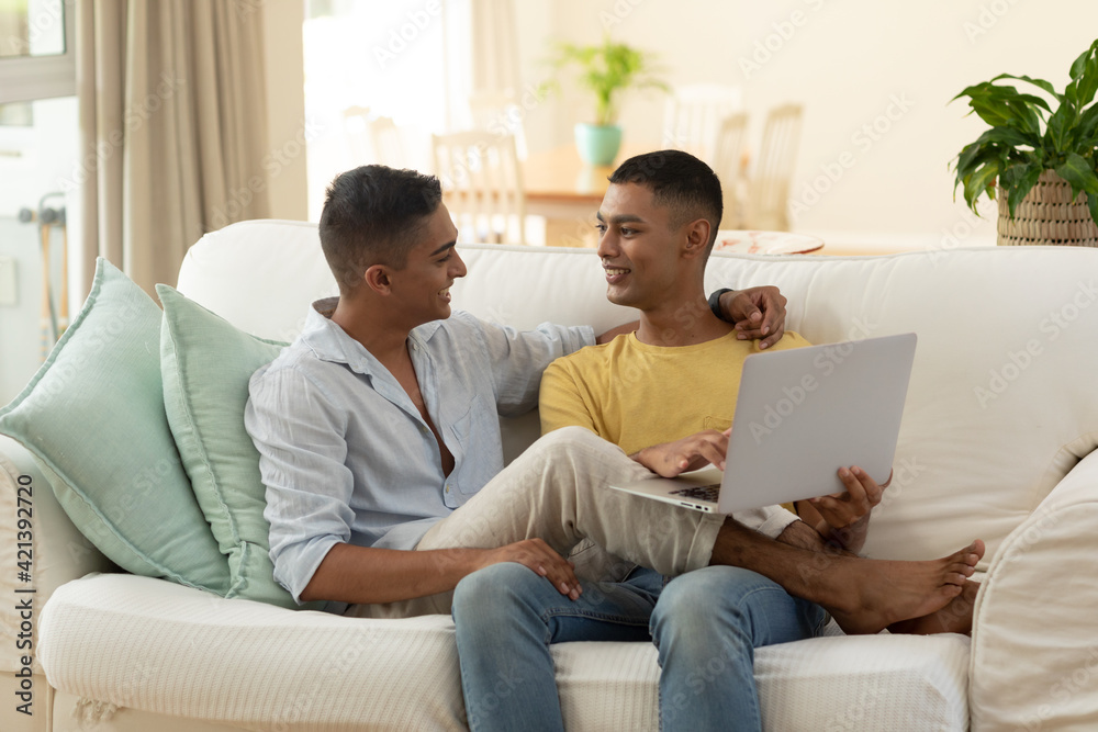 Diverse gay male couple sitting on sofa using laptop computer and embracing Diverse gay male couple sitting on sofa using laptop computer and embracing
