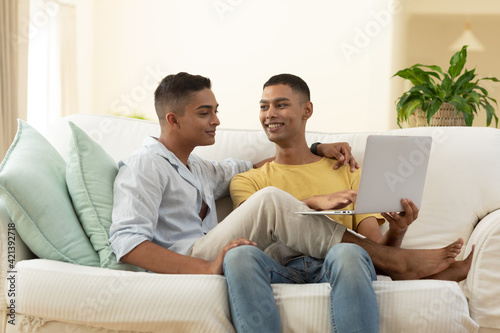 Diverse gay male couple sitting on sofa using laptop computer and embracing