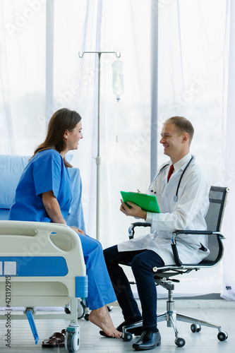 Woman caucasian patient sitting on bed and caucasian doctor sitting on chair are talking, both are smiling look happy. Doctor man carrying stethoscope on neck and hold writing on note in office room