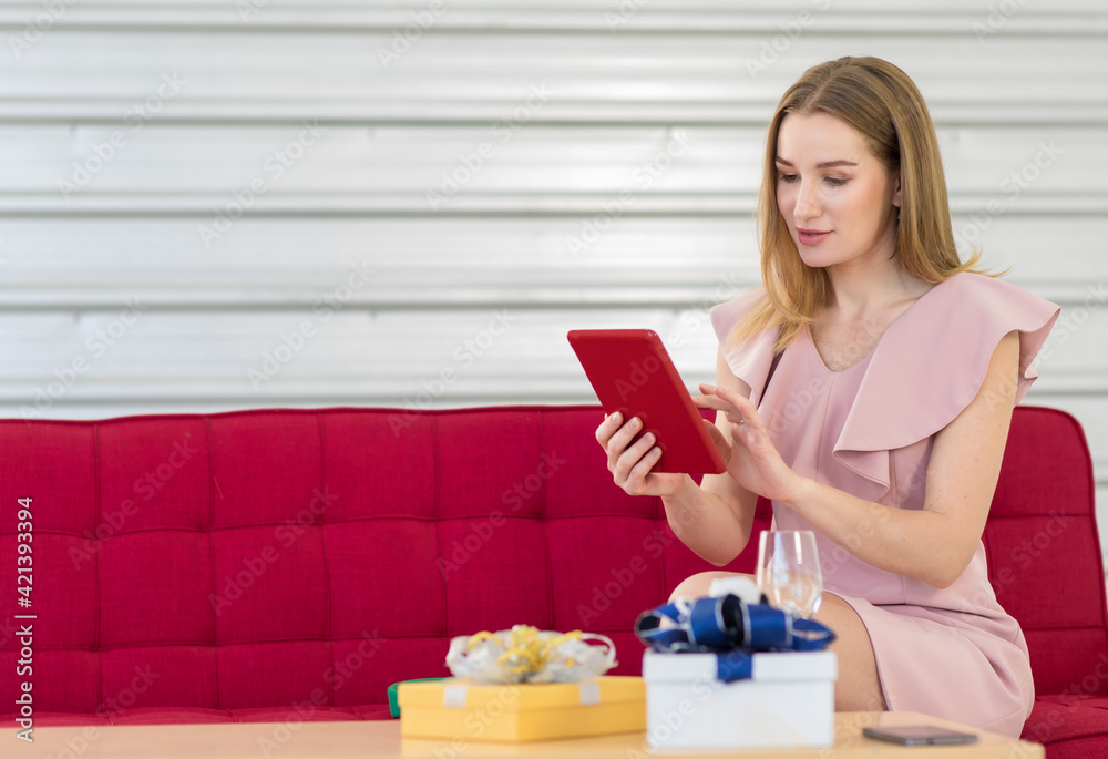 Focus to adult caucasian woman blonde hair and pink dress look online seller sitting at red sofa chair holding touch and look at tablet with gift box, smartphone and glass on table in blur foreground Focus to adult caucasian woman blonde hair and pink dress look online seller sitting at red sofa chair holding touch and look at tablet with gift box, smartphone and glass on table in blur foreground