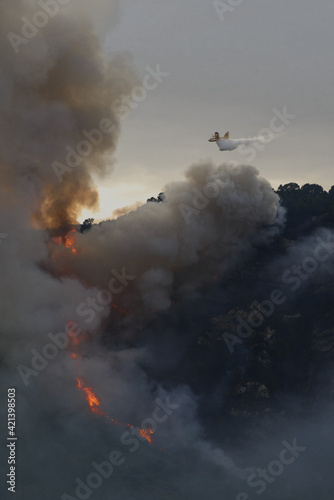 big fire in the mountains with high flames and a long tongue of fire between big clouds of smoke and a firefighting plane dumps water on the fire to try to tame it