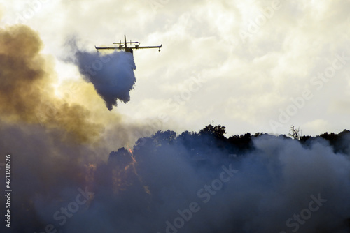 big fire in the mountains with high flames and a long tongue of fire between big clouds of smoke and a firefighting plane dumps water on the fire to try to tame it