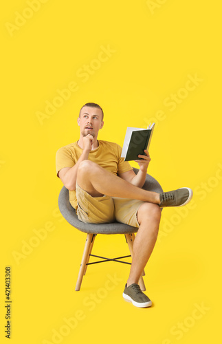 Young man reading book while sitting in armchair on color background