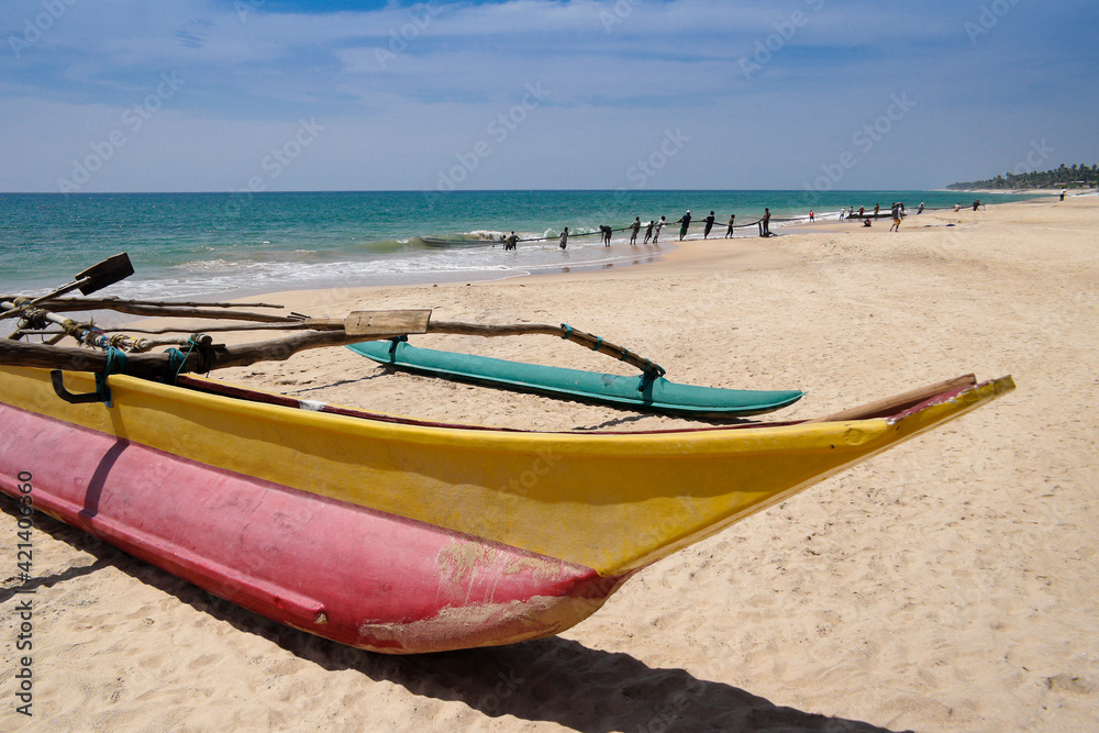 At Ambalangoda, Sri Lanka, a traditional outrigger sea canoe (oru) rests on the sand as fishermen in the background haul in their large net (madela). At Ambalangoda, Sri Lanka, a traditional outrigger sea canoe (oru) rests on the sand as fishermen in the background haul in their large net (madela).
