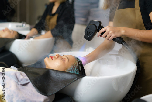 Hairdresser is using Nano Mist Hair Treatment Streamer Machine, Modern equipment for Young Happy Asian Beautiful Caucasian Woman Customer on Shampoo Chair In Hair Salon. Beauty Concept..