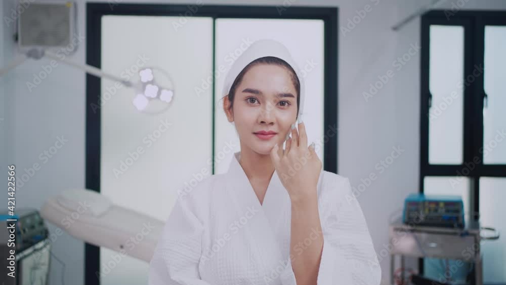 Asian woman in bathrobe and towel on her head gently touching skin on her face and smiling to be satisfied with herself a beauty while looking at camera like a mirror in beauty clinic's operating room Asian woman in bathrobe and towel on her head gently touching skin on her face and smiling to be satisfied with herself a beauty while looking at camera like a mirror in beauty clinic's operating room