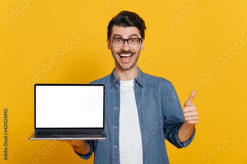 Amazed excited cheerful caucasian unshaven guy in glasses and denim shirt holds open laptop with blank white screen, showing thumbs up gesture, standing on isolated orange background, friendly smiling