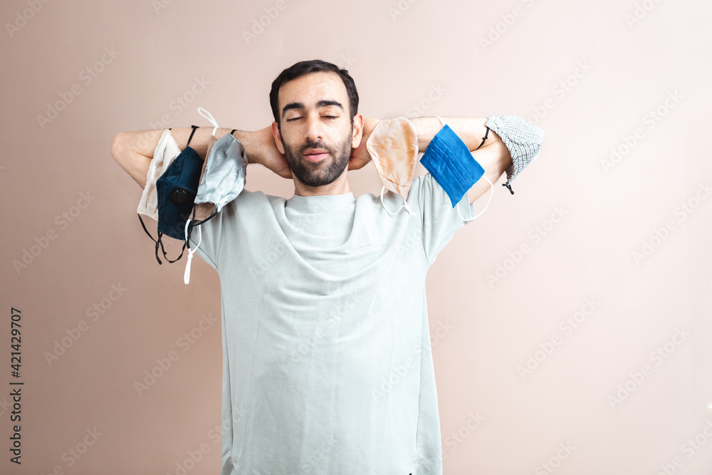 Young middle eastern adult man holding many face masks on the arms. Wearing plain t-shirt. Creative studio shot on nude pastel backdrop. Closeup, space for text Young middle eastern adult man holding many face masks on the arms. Wearing plain t-shirt. Creative studio shot on nude pastel backdrop. Closeup, space for text