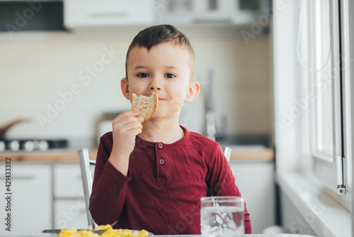 A child in the afternoon on a white light kitchen in a burgundy sweater eats an omelet