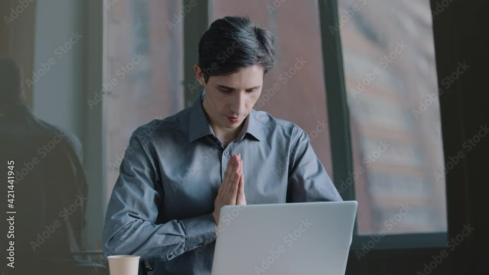 Portrait of caucasian business man manager wears formal shirt sitting at table looking into laptop folds palms hands in supplication pray expresses gratitude tilting head greets in chinese tradition Portrait of caucasian business man manager wears formal shirt sitting at table looking into laptop folds palms hands in supplication pray expresses gratitude tilting head greets in chinese tradition