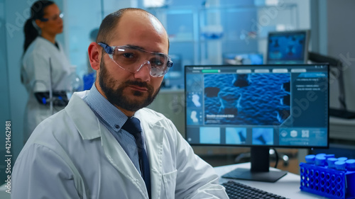 Overworked scientist with protection glasses looking at camera sighing sitting in research laboratory. Doctors examining virus evolution using high tech and chemistry tools for medical research.