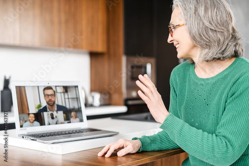 Serious mature lady grandmother teacher sitting at kitchen table, waving at laptop with people profiles, talking with webinar conference participants online, having virtual meeting lesson via laptop