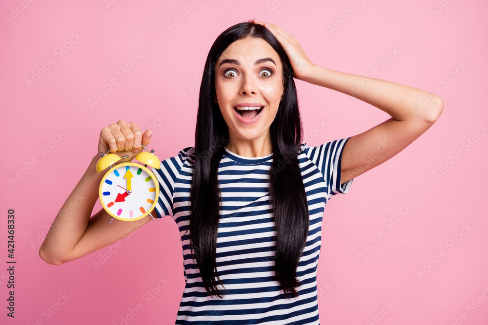 Photo portrait of amazed woman with open mouth touching head holding yellow clock in one hand isolated on pastel pink colored background Photo portrait of amazed woman with open mouth touching head holding yellow clock in one hand isolated on pastel pink colored background