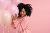 Happy pleased African woman with curly hair holding a lot of balloons, enjoys cool party, wears pink sweater, celebrates birthday, looking at camera, isolated on studio pink background. Festive event