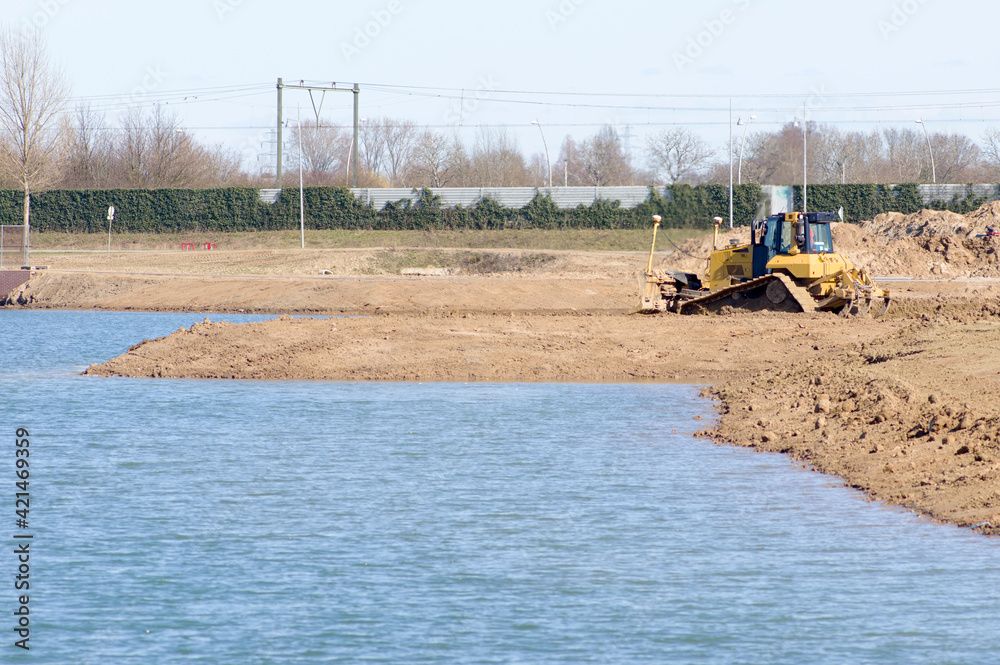 Large yellow bulldozer is moving ground at a quay of a lake with a railway in the background
