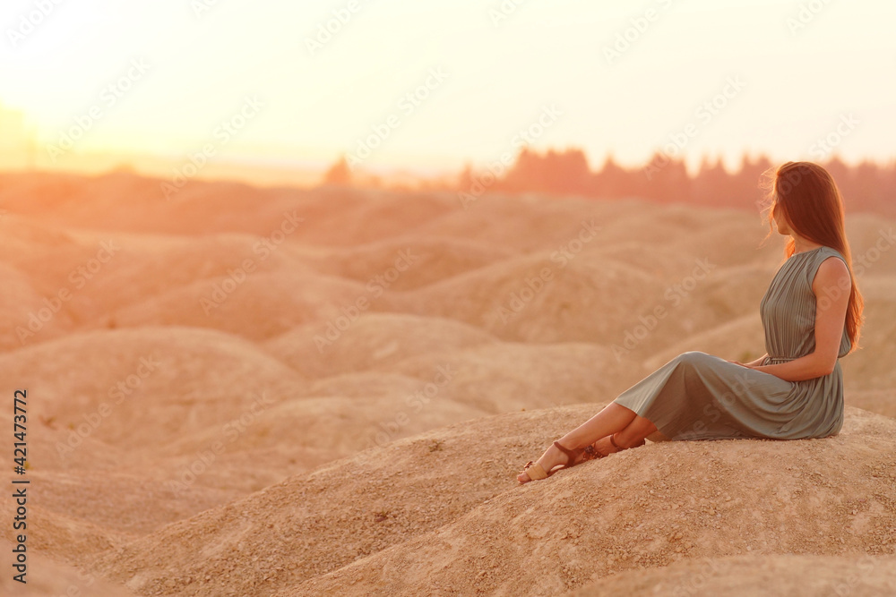 Young beautiful woman with long hair in blue dress sitting elegant and thoughtful on sand at sunrise in sandy desert