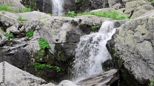 Mountain spring stream flows among gray rocks. Waterfall cascades down from cliffs. Water foams and splashes. Green grass grows on boulders on shore.