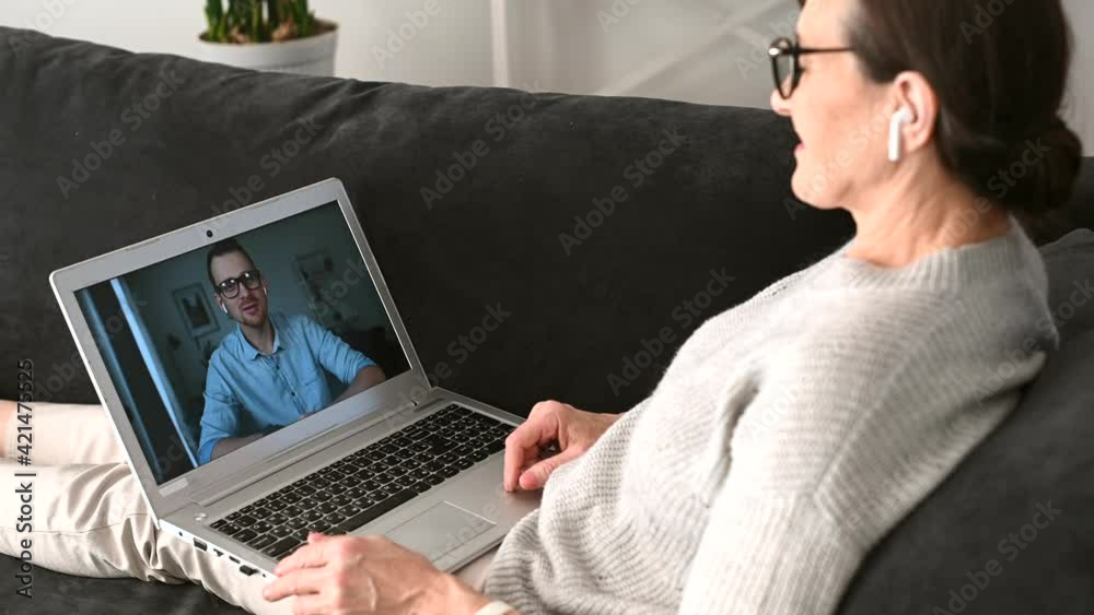 A smiling senior woman is sitting in the living room, relaxing and talking to her adult son at the laptop screen via video call A smiling senior woman is sitting in the living room, relaxing and talking to her adult son at the laptop screen via video call