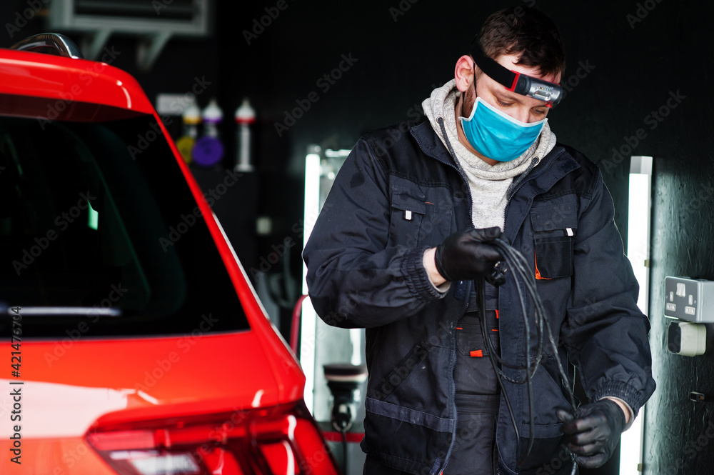 Naklejka premium Car detailing concept. Man in face mask with orbital polisher in repair shop polishing orange suv car.