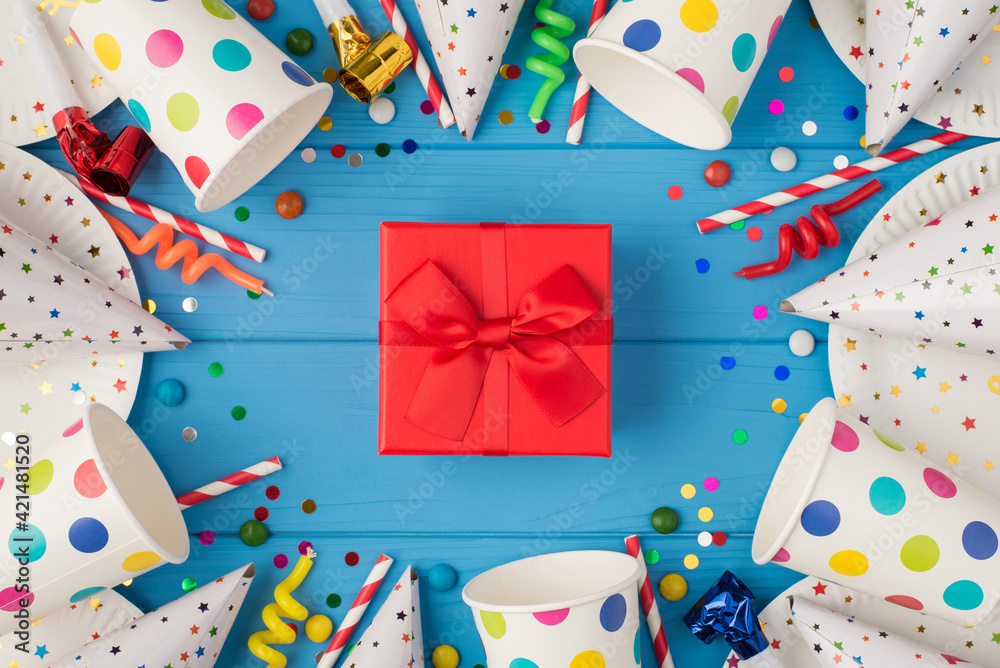 Top view photo of red gift box with ribbon bow in the middle of birthday composition and party accessories on isolated blue wooden table background Top view photo of red gift box with ribbon bow in the middle of birthday composition and party accessories on isolated blue wooden table background