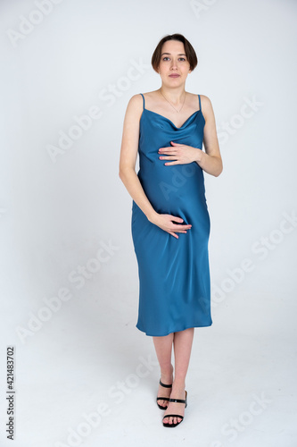 Studio portrait of young adult pregnant woman in blue dress on white gray background, happy pregnancy concept