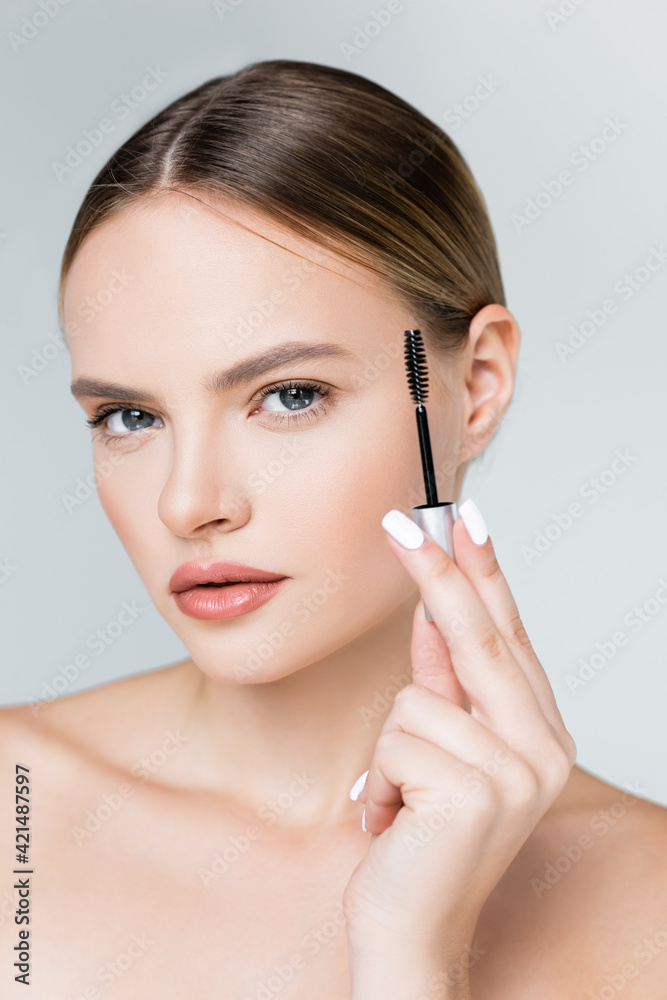 young woman holding mascara and looking at camera isolated on grey