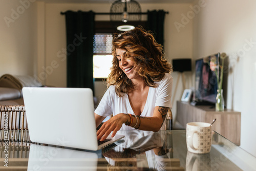 Portrait of a cheerful woman at home with a laptop and a cup of coffee on the table. 30 year old girl connected online to a meeting with friends and family. Telework concept