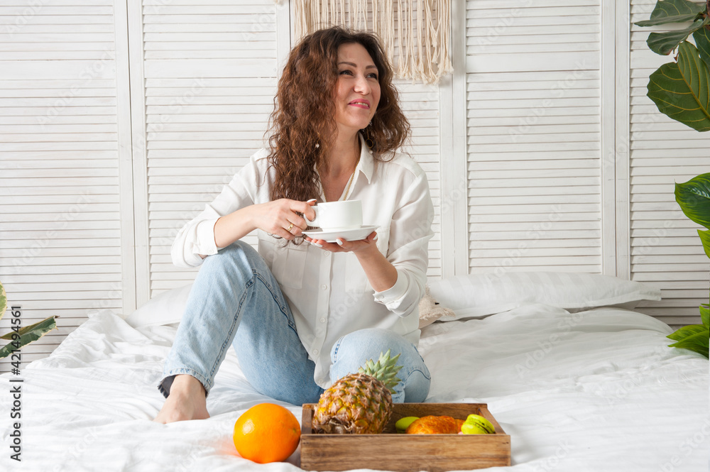 Brunette woman in white clothes is drinking morning coffee with cookies and fruits in bed