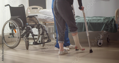 Cropped shot of doctor helping patient with bandaged leg walking on crutches in hospital ward