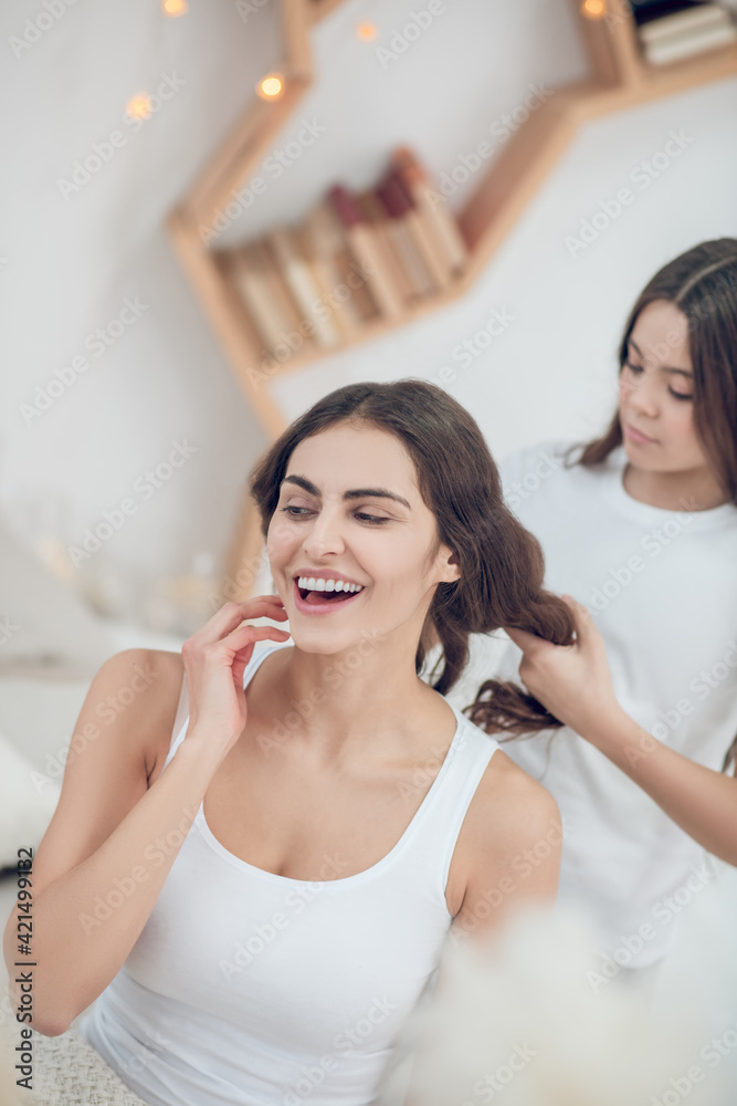 Girl weaving braid to emotional woman in pajamas Girl weaving braid to emotional woman in pajamas