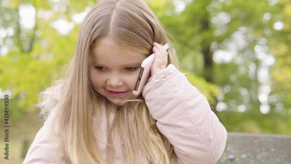 Cute little Caucasian girl talks on a smartphone with a smile as she sits on a bench in a park - closeup Cute little Caucasian girl talks on a smartphone with a smile as she sits on a bench in a park - closeup