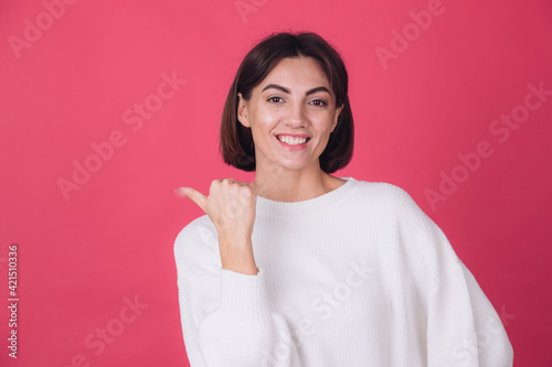 Beautiful woman in casual white sweater on pink red background smiling point left with thumb finger isolated copy space