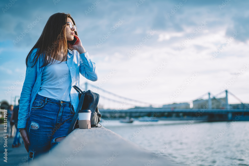 Attractive Caucaisan woman in spectacles using cellphone internet connection for communicate in roaming, millennial female tourist making international mobile conversation thinking at embankment