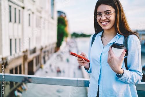 Half length of cheerful woman in trendy spectacles smiling at camera spending free time outdoors using mobile phone,millennial hipster girl with smartphone technology and coffee to go laughing in city