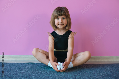 Little gymnast girl with blond hair in a beautiful black leotard is sitting on a background of a pink wall photo