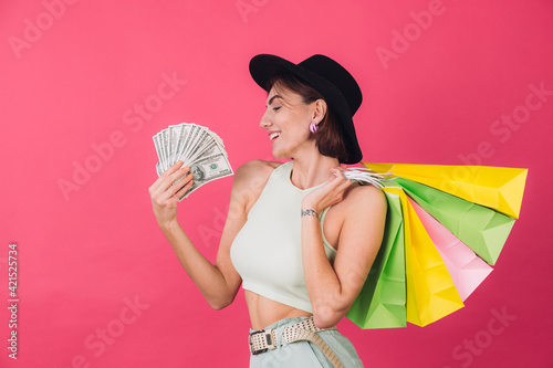 Stylish woman in hat on pink red background isolated space smile excited hold fan of 100 dollars money and shopping bags, happy positive smiling