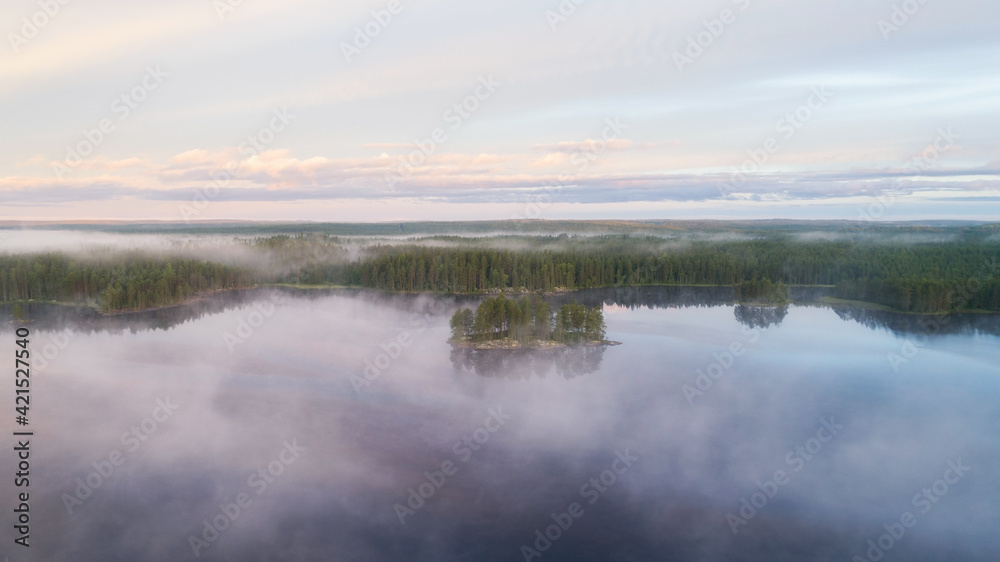 Island with pine trees in morning fog on lake at sunrise. Mysterious island. Atmospheric sunrise over summer forest lake.