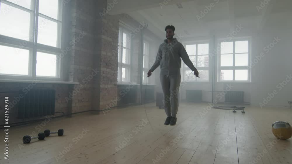 Slow-motion full shot of young african-american sportsman in grey tracksuit skipping rope while training alone in smoky loft room