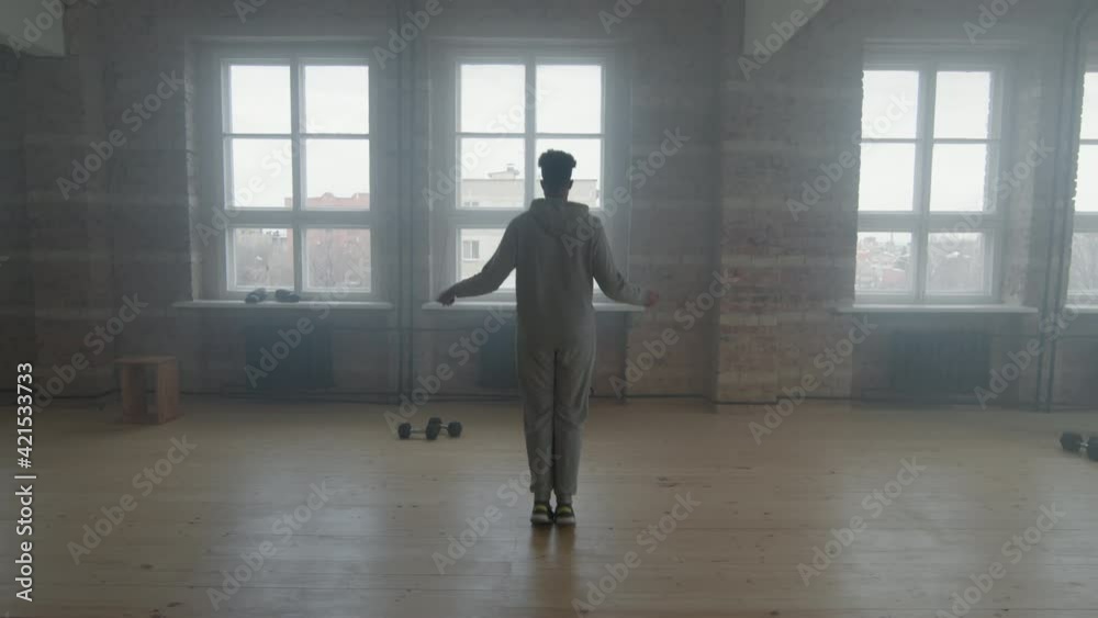 Rear-view full shot with slowmo of young sportsman in grey tracksuit jumping rope in front of window in smoky loft style room alone Rear-view full shot with slowmo of young sportsman in grey tracksuit jumping rope in front of window in smoky loft style room alone