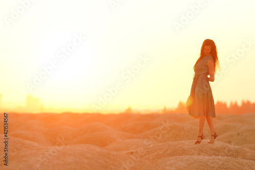 Beautiful woman with long hair in blue dress with straw hat in hand standing on sand in desert at sunrise, backlight