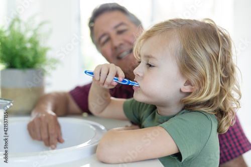 Smiling caucasian grandfather in bathroom kneeling beside grandson brushing teeth