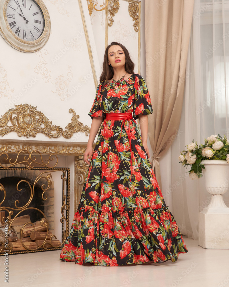 Pretty elegant woman in colorful vivid dress with flower design, sleeves and red belt standing and posing in bright interior
