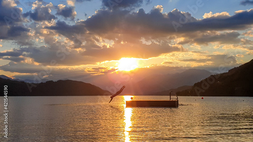 A man jumping into the Millstaettersee lake from a wooden platform during the sunset. The sun is setting behind high Alps. Calm surface of the lake reflects the orange sky and the mountains. Serenity photo