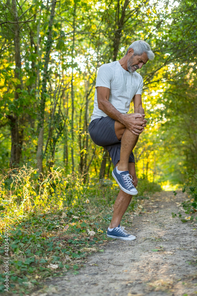 Mature man suffering from knee pain while standing in forest Mature man suffering from knee pain while standing in forest