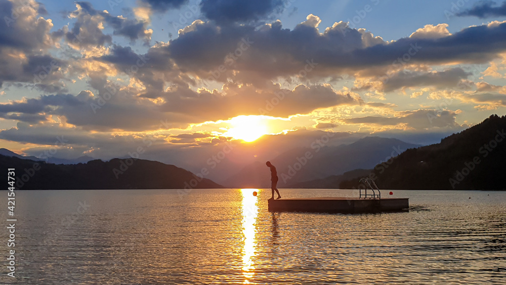 A man running along wooden platform drifting on the MIllstaetter lake during the sunset. Jumping into deep water. The sun sets behind high Alps. Calm surface of the lake reflects the orange sky. Fun