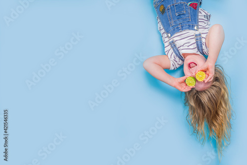 Slika na platnu Little smiling blond girl upside down fluttering hair holding halves of fresh lemon and lime fruit near eyes, bright blue wall