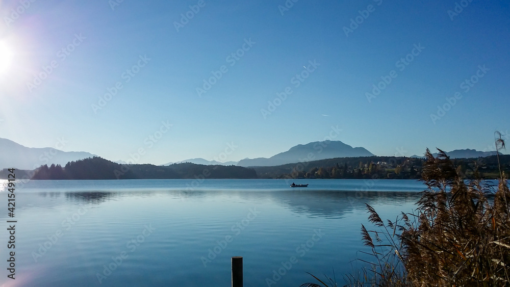 A panoramic view on the Faaker lake in Austrian Alps. The lake is surrounded by high mountains. Calm surface of the lake reflects the surrounding. Sweet flag at the shore. Clear blue sky. Serenity