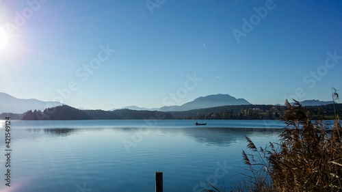 A panoramic view on the Faaker lake in Austrian Alps. The lake is surrounded by high mountains. Calm surface of the lake reflects the surrounding. Sweet flag at the shore. Clear blue sky. Serenity