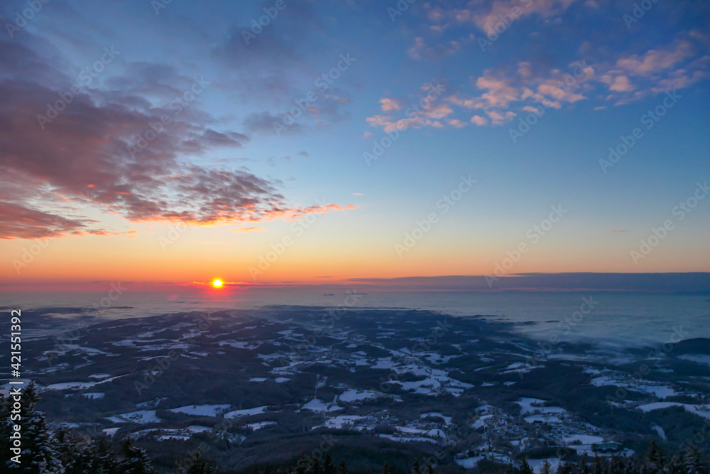 A sunrise seen from snow-capped peak of Schoeckl in Austrian Alps. The sky is bursting with orange and pink. Distant view on Graz, shrouded with fog. Winter wonderland. Day break. Calmness
